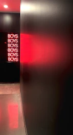 A dimly lit hallway features a black wall reflecting a vivid red neon sign that repeatedly spells out 'BOYS'. The illumination casts a red glow across the floor and wall, creating a modern and slightly mysterious atmosphere.