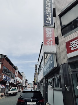 A narrow urban street lined with commercial buildings, featuring several signs in Korean. Vehicles, including a black car in the foreground, are parked along the sides. The sky is partly cloudy, and the buildings have a mix of modern and traditional architectural elements with brick and metal facades.