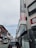 A narrow urban street lined with commercial buildings, featuring several signs in Korean. Vehicles, including a black car in the foreground, are parked along the sides. The sky is partly cloudy, and the buildings have a mix of modern and traditional architectural elements with brick and metal facades.