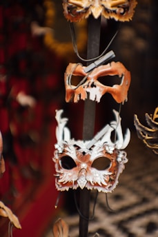 The image shows a display of intricately designed masks hanging on a vertical rod. The masks feature various themes, including a skull motif and a detailed, feathered design with horns. The background is blurred but displays a patterned floor and dark surroundings possibly indicative of a shop or display area.