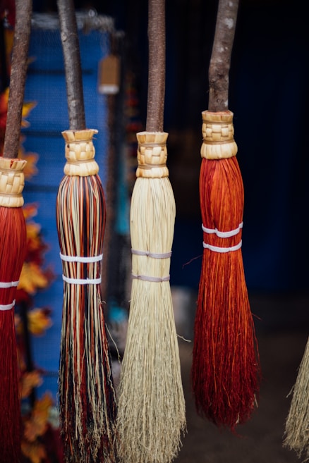 Four handmade brooms with wooden handles and straw-based bristles hang side by side. The bristles vary in color, showcasing shades of red, beige, and mixed patterns. They are tied at the base with simple white strings, and the texture of the straw gives a rustic appearance. A blurred background features hints of blue and other colors, suggesting a market setting.