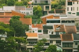 A cluster of residential houses with various architectural styles, featuring orange and brown tiled roofs amidst lush greenery. The mix of modern and traditional structures creates a scenic urban landscape.