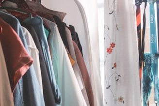 A collection of clothes hanging on a rack next to a white curtain with floral embroidery. The clothes are of various colors, including shirts and jackets, and hangers of different colors are visible. Next to the clothes, lanyards with text are hanging, adding a splash of color.