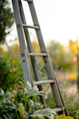 An adjustable aluminum telescopic ladder extended against a garden wall with lush greenery in the background.