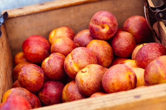 A wooden crate filled with numerous ripe nectarines, showcasing their reddish and orange hues. The texture of the fruit is smooth, with some speckled variations. The crate is simple and rustic, emphasizing a natural and fresh presentation.