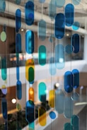 Colored glass panels or hanging decorative pieces in various shapes and hues of blue, green, and yellow are suspended in an indoor setting with a blurred background of lights, plants, and modern architectural elements.