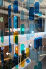 Colored glass panels or hanging decorative pieces in various shapes and hues of blue, green, and yellow are suspended in an indoor setting with a blurred background of lights, plants, and modern architectural elements.