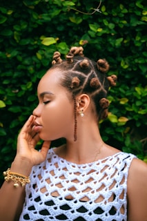 A person with a unique hairstyle featuring intricately styled hair buns and braids is seen in profile. The individual is wearing a white crochet top and accessorized with gold jewelry including earrings and a bracelet. The background consists of vibrant green foliage.