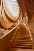 A grand and elegantly designed interior space featuring a large, wooden staircase that ascends towards a ceiling adorned with a modern, geometric glass skylight. The architecture showcases flowing, organic curves and warm, wooden textures, creating an inviting atmosphere.