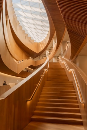 A grand and elegantly designed interior space featuring a large, wooden staircase that ascends towards a ceiling adorned with a modern, geometric glass skylight. The architecture showcases flowing, organic curves and warm, wooden textures, creating an inviting atmosphere.