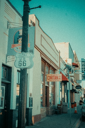 A vintage-style image of a street lined with retro buildings, including signs for Route 66 and Civitan on 66. The architecture features large windows and a mix of metal and brick materials. A light post stands in the foreground with the iconic US 66 shield. The scene suggests a small-town American street, hinting at mid-20th century design.