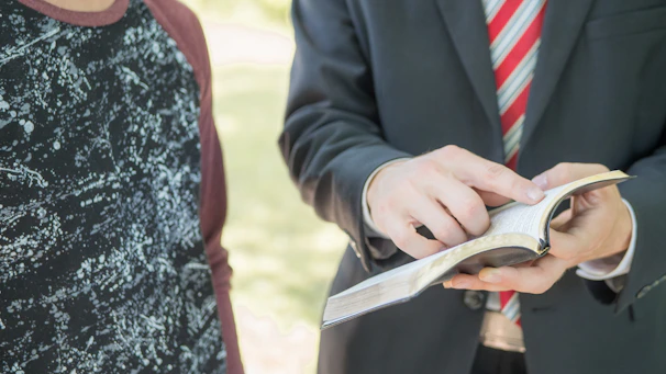 Two people are interacting. One person is holding and pointing at a book, possibly interacting or explaining something in it. The person pointing is wearing a suit with a striped tie, suggesting formality. The other person is dressed casually in a patterned shirt.