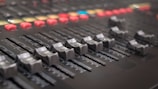 A close-up view of a control panel or mixing console with multiple sliders and knobs. The focus is on several horizontal sliders used for adjusting audio levels, with numerous buttons and lights in the background enhancing its technological appearance.