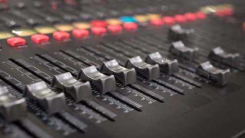 A close-up view of a control panel or mixing console with multiple sliders and knobs. The focus is on several horizontal sliders used for adjusting audio levels, with numerous buttons and lights in the background enhancing its technological appearance.