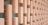A series of vertically oriented, light-colored wooden blocks arranged in a staggered, grid-like pattern. Each block is connected to the next by thin horizontal rods, creating a three-dimensional effect. The backdrop is a muted green wall, providing contrast to the wood texture.