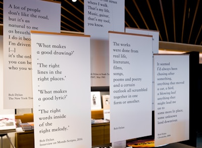 Several large posters are hung in a room, each displaying quotes from Bob Dylan. The quotes cover topics like creativity, life experiences, and personal reflection. The room has a modern design with visible wooden elements on the ceiling and a few pieces of furniture and books in the background.