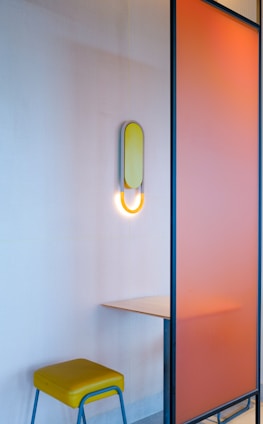 A minimalist interior setting with a small wooden table attached to a vibrant orange partition. A yellow wall lamp with a circular design illuminates the space softly. A yellow cushioned stool with light blue legs is placed under the table.