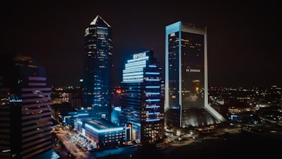 An urban nightscape featuring illuminated skyscrapers with bright neon lights and reflective glass surfaces. The city is alive with glowing signs and a network of light trails from streets below, set against a dark night sky.