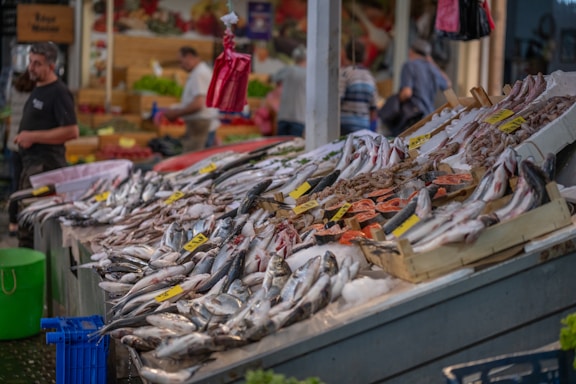 A busy fish market stall displaying a variety of fresh seafood including fish and shrimp on ice. The setup includes yellow price tags, and there are blurred figures of people in the background, indicating a lively market atmosphere.