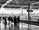 A group of people is waiting on a spacious, modern railway platform. A high-speed train is visible on the adjacent track, with the city skyline and overhead electric lines in the background. The scene is set under an architectural canopy with a geometric design, creating a structural and organized atmosphere.