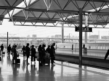 A group of people is waiting on a spacious, modern railway platform. A high-speed train is visible on the adjacent track, with the city skyline and overhead electric lines in the background. The scene is set under an architectural canopy with a geometric design, creating a structural and organized atmosphere.