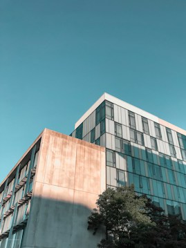 A modern building with a minimalist design, featuring large glass windows reflecting the sky. The structure is partly shaded, with some sunlight creating a warm tone on the facade. A few trees are visible at the base, adding a touch of nature to the architectural scene.