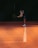 A person in athletic attire is on a tennis court, bending slightly with a racket in hand, preparing to play. They are wearing a cap, tank top, and shorts, and the court has a warm, orange hue with a white line running through the center.