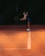 A person in athletic attire is on a tennis court, bending slightly with a racket in hand, preparing to play. They are wearing a cap, tank top, and shorts, and the court has a warm, orange hue with a white line running through the center.