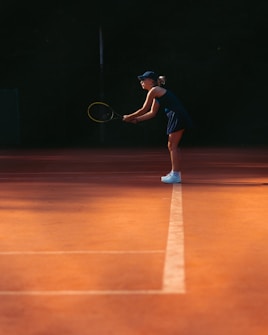 A person in athletic attire is on a tennis court, bending slightly with a racket in hand, preparing to play. They are wearing a cap, tank top, and shorts, and the court has a warm, orange hue with a white line running through the center.