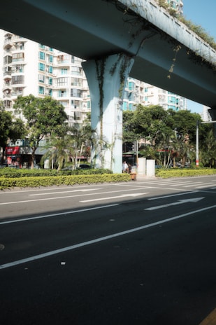 A modern urban scene with a concrete overpass draped in foliage, set against a backdrop of residential high-rise buildings. The street below is empty, bordered by well-maintained bushes and tall trees that add greenery to the urban landscape.