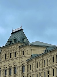 A historic building with ornate architectural details, including arched windows and intricate moldings. The roof features a steep, pitched design with decorative elements along its edge, and the structure is primarily beige with green roofing.