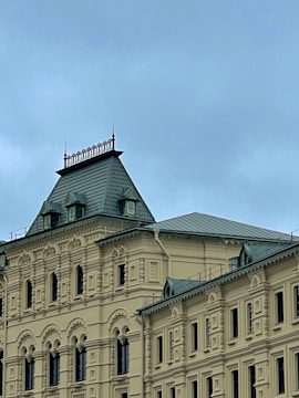 A historic building with ornate architectural details, including arched windows and intricate moldings. The roof features a steep, pitched design with decorative elements along its edge, and the structure is primarily beige with green roofing.