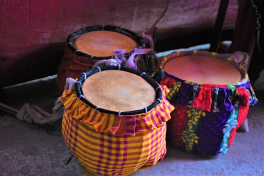 Three traditional drums are placed on the ground, each covered in colorful, patterned fabrics. The fabrics feature a mix of vibrant hues and designs, adding a cultural and artistic touch to the scene. The drums are positioned closely together, suggesting a ready-for-performance setup.