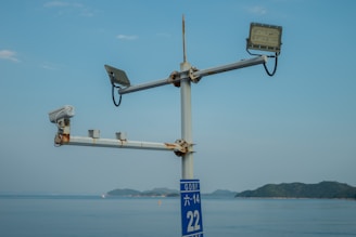 A metal structure with a CCTV camera and two floodlights is mounted on a pole, overlooking a body of water with distant islands in the background. The sky is clear with a few clouds.