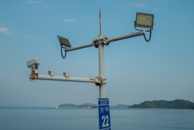 A metal structure with a CCTV camera and two floodlights is mounted on a pole, overlooking a body of water with distant islands in the background. The sky is clear with a few clouds.