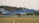 An airplane with KLM branding is landing on a runway, surrounded by green grass and trees in the background. Smoke can be seen from the landing gear touching the runway, and a small building is visible behind the trees.