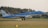 An airplane with KLM branding is landing on a runway, surrounded by green grass and trees in the background. Smoke can be seen from the landing gear touching the runway, and a small building is visible behind the trees.