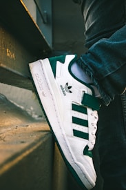 A close-up view of a person wearing white and green sports shoes, with the foot resting on a concrete ledge. The sneaker features a branded logo and signature stripes. The scene includes part of the leg wearing dark blue jeans, set against the background of some steps and a wall.