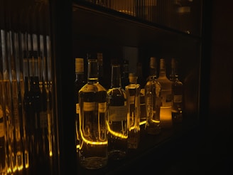 A collection of various liquor bottles are displayed on a dimly lit shelf, with warm golden lighting creating reflections and highlighting the bottles' shapes.