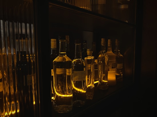 A collection of various liquor bottles are displayed on a dimly lit shelf, with warm golden lighting creating reflections and highlighting the bottles' shapes.
