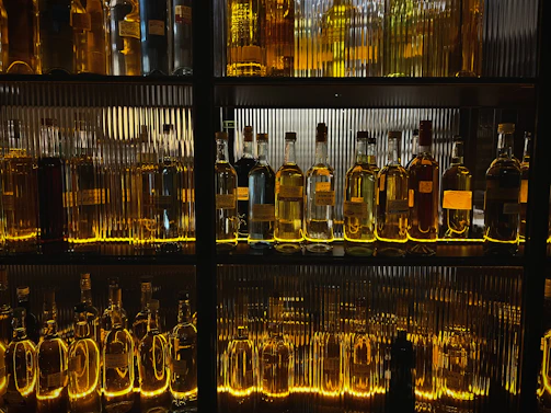 A rustic wooden shelf lined with an array of colorful boutique spirit bottles, softly lit to highlight their unique labels.