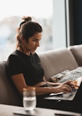 A woman is sitting on a couch and working on a laptop. She is wearing a black shirt and appears focused on her work. A glass of water is placed on a table in the foreground. The setting seems to be a cozy, well-lit living space with large windows.