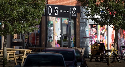 A street scene featuring a café with a black and white sign above the entrance displaying contact information. Large trees and a car partially obstruct the view. The storefront has colorful posters and graffiti art around, with a bench under the trees. A person wearing a white helmet and a yellow jacket is sitting at a picnic table next to a bicycle.