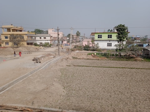A semi-urban landscape with several two-story buildings and open plots of land. The sky is overcast, and the ground appears dry with patches of greenery. People are walking along a dirt road, and there is some construction material scattered around.