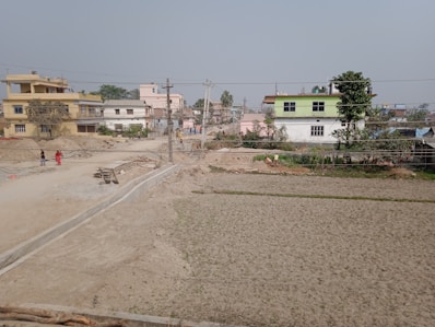 A semi-urban landscape with several two-story buildings and open plots of land. The sky is overcast, and the ground appears dry with patches of greenery. People are walking along a dirt road, and there is some construction material scattered around.