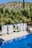 A serene outdoor setting features a clear, blue swimming pool surrounded by lush green palm trees. Cabana-style day beds with white drapes are positioned alongside the pool, offering a luxurious and relaxing atmosphere. In the background, a hilly landscape with sparse greenery and a bright blue sky is visible.