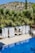 A serene outdoor setting features a clear, blue swimming pool surrounded by lush green palm trees. Cabana-style day beds with white drapes are positioned alongside the pool, offering a luxurious and relaxing atmosphere. In the background, a hilly landscape with sparse greenery and a bright blue sky is visible.
