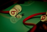 A pair of Beats in-ear headphones with red cables and white buds are placed on a reflective surface, likely a device with a shiny green cover, alongside an Apple logo.