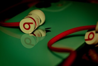 A pair of Beats in-ear headphones with red cables and white buds are placed on a reflective surface, likely a device with a shiny green cover, alongside an Apple logo.