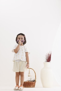 A young child wearing a light-colored outfit stands indoors, holding sunglasses to their face. Beside them is a wicker basket filled with dried plants, and a large white vase also containing dried stems. The setting is minimalist with neutral colors.
